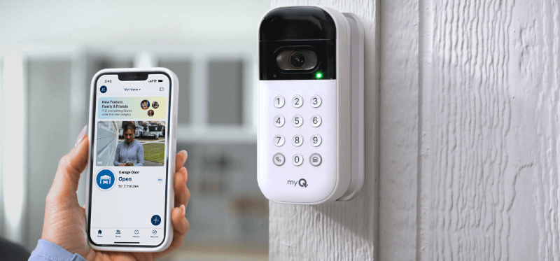 In a sunny, inviting room, a hand holds up a smartphone while navigating a cleanly designed app, sending a command for a garage door to open. Nearby is a modern white and black myQ keypad showing a little green light of affirmation.