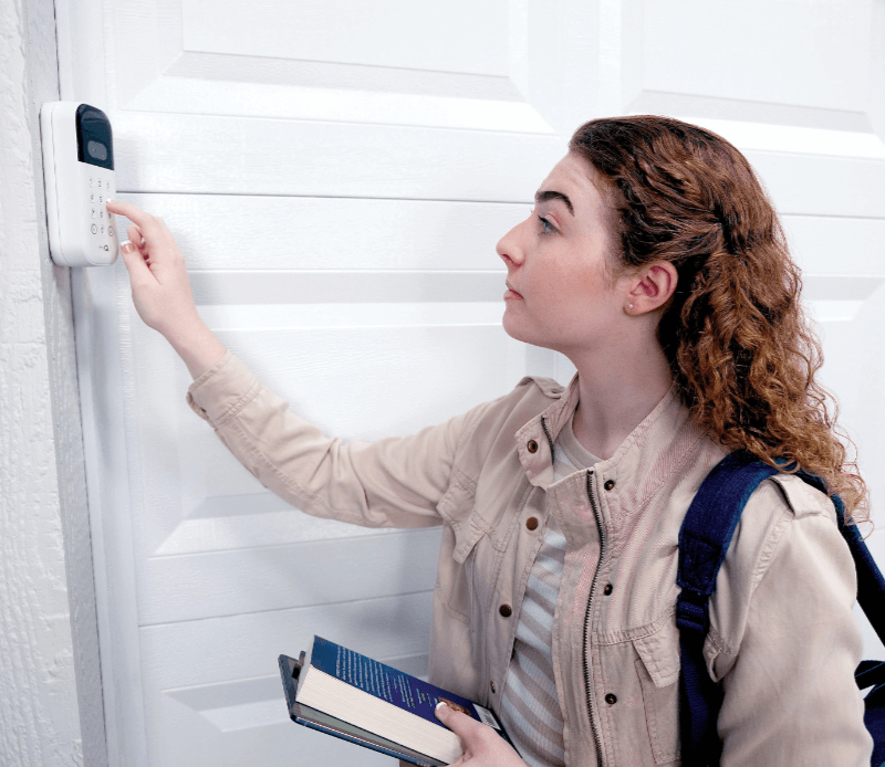 A young person with a backpack and books enters a code into their MyQ smart garage door opener keypad in front of their garage door.